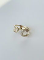 Create a professional, high-quality product photo using the exact same ring from the reference image, without changing its design, shape, or details. The image should feature the same gold-plated safety pin–style ring with its real textures, stones, and structure. Use soft natural lighting, a clean white or light neutral background, sharp focus on the ring, and a modern, minimalistic, luxurious aesthetic. Maintain realism and ensure the ring looks identical to the original item.