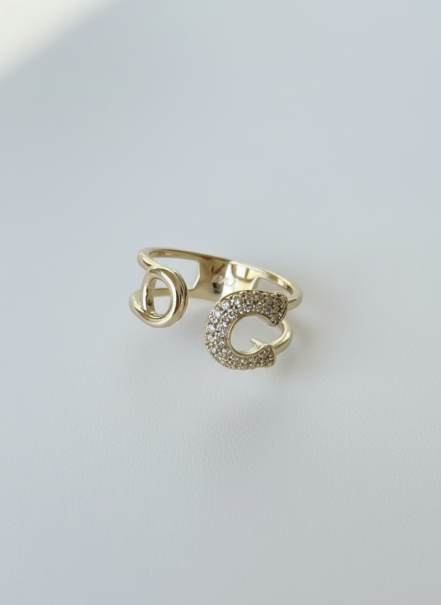 Create a professional, high-quality product photo using the exact same ring from the reference image, without changing its design, shape, or details. The image should feature the same gold-plated safety pin–style ring with its real textures, stones, and structure. Use soft natural lighting, a clean white or light neutral background, sharp focus on the ring, and a modern, minimalistic, luxurious aesthetic. Maintain realism and ensure the ring looks identical to the original item.